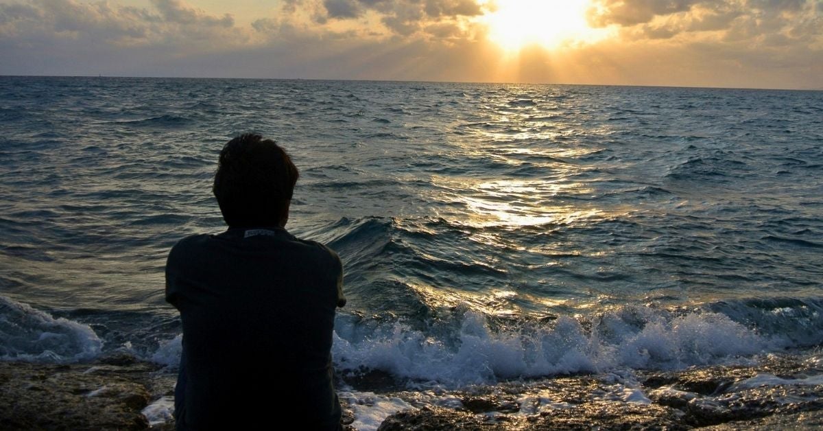 A person sits on rocky shore, facing a tranquil ocean under a cloudy sky. The golden sunset beams create a reflective, serene atmosphere.