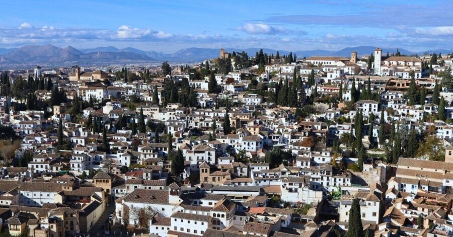 Aerial view of a historic Santander, Spain. White buildings with terracotta roofs nestled among tall cypress trees cover the landscape as mountains rise in the background under a blue sky.