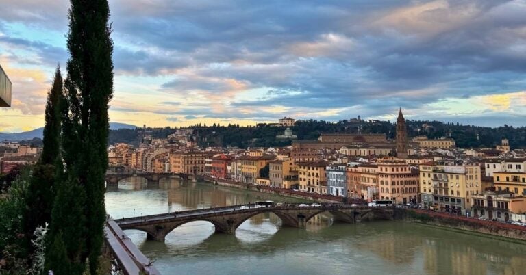 View of Florence, Italy, featuring the Arno River, a stone bridge, cypress trees, and historic buildings under a cloudy sky.