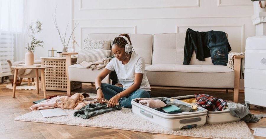 A woman wearing headphones sits on a carpeted floor, packing clothes into an open suitcase. The room has a cozy, tidy feel with natural light.