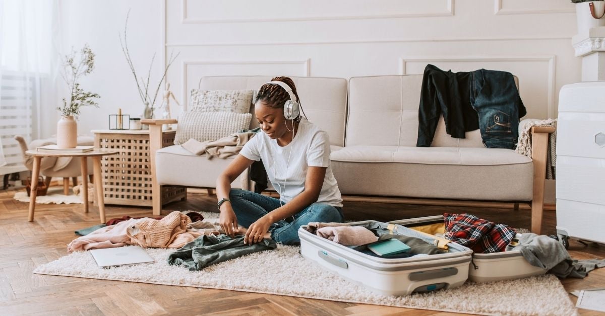 A woman wearing headphones sits on a carpeted floor, packing clothes into an open suitcase. The room has a cozy, tidy feel with natural light.