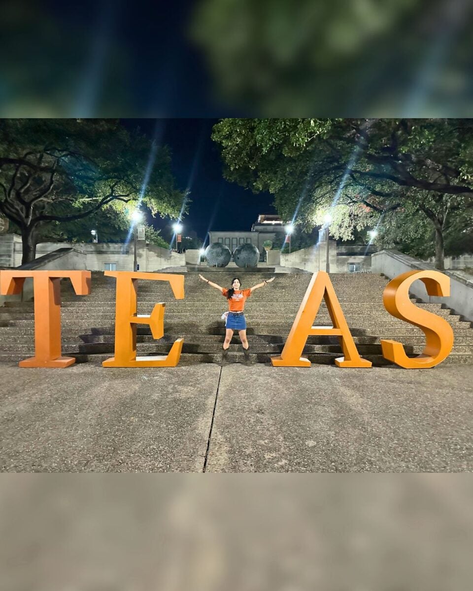 A person joyfully stands with arms raised between large, illuminated orange letters spelling "TEXAS" on stone steps at night, under trees and lampposts.