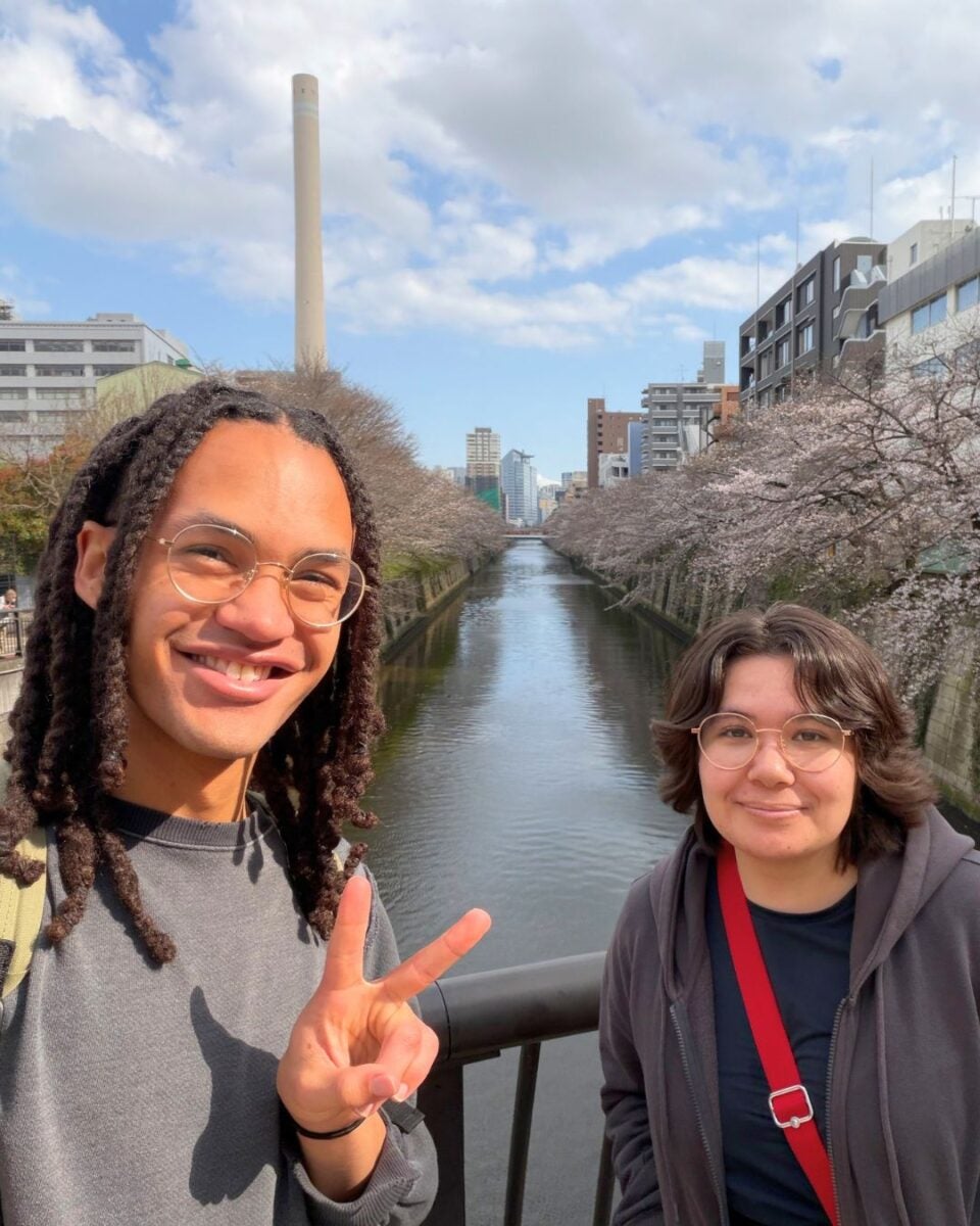 Two people smiling on a bridge with cherry blossoms, a river, and city buildings in the background. One person flashes a peace sign. Bright, sunny day.