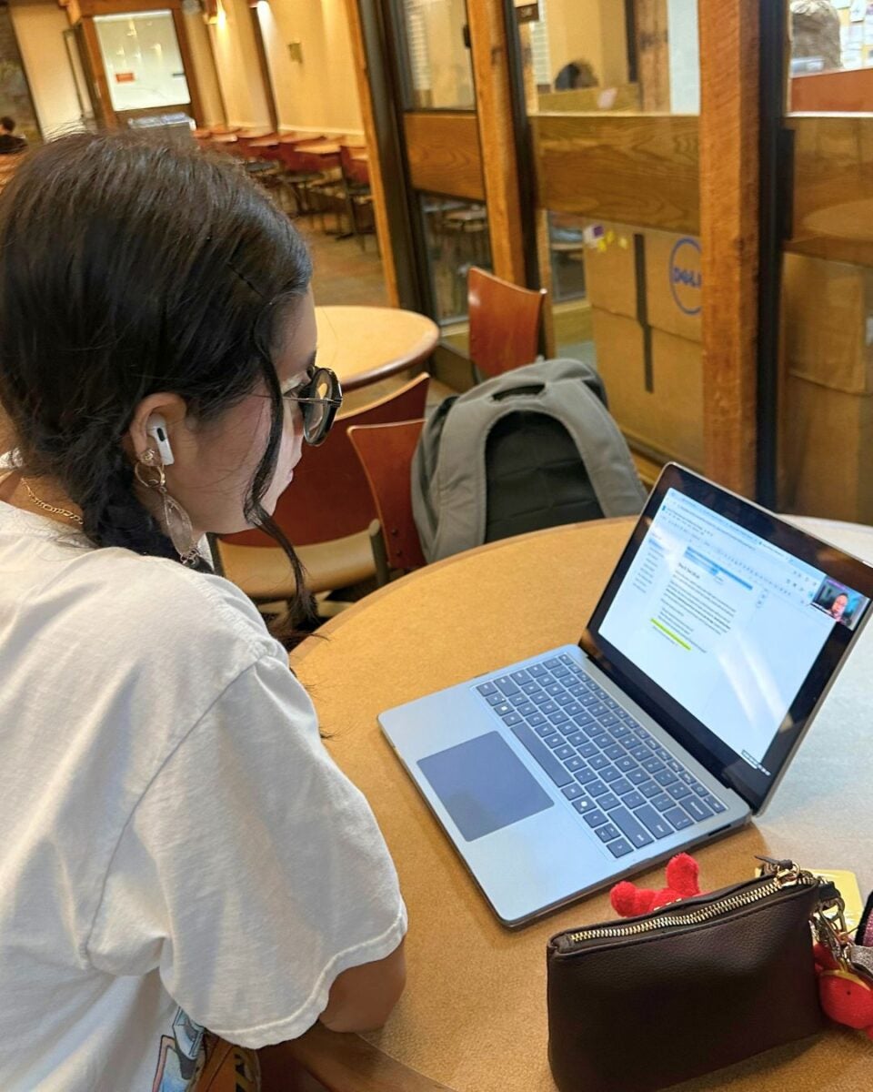 A person with earbuds studies on a laptop at a round table in a cafe. They are focused, surrounded by empty chairs, with a backpack nearby.
