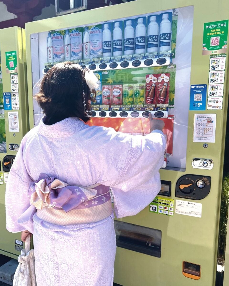 A person in a lavender kimono uses a vending machine stocked with various drinks.