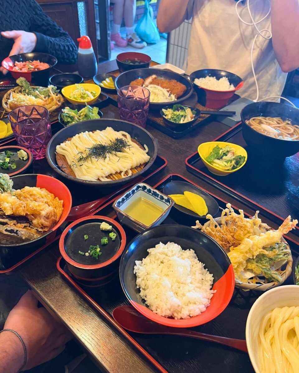 A table filled with assorted Japanese dishes, including rice, tempura, noodles, and pickles. Two people sit nearby, creating a cozy dining vibe.
