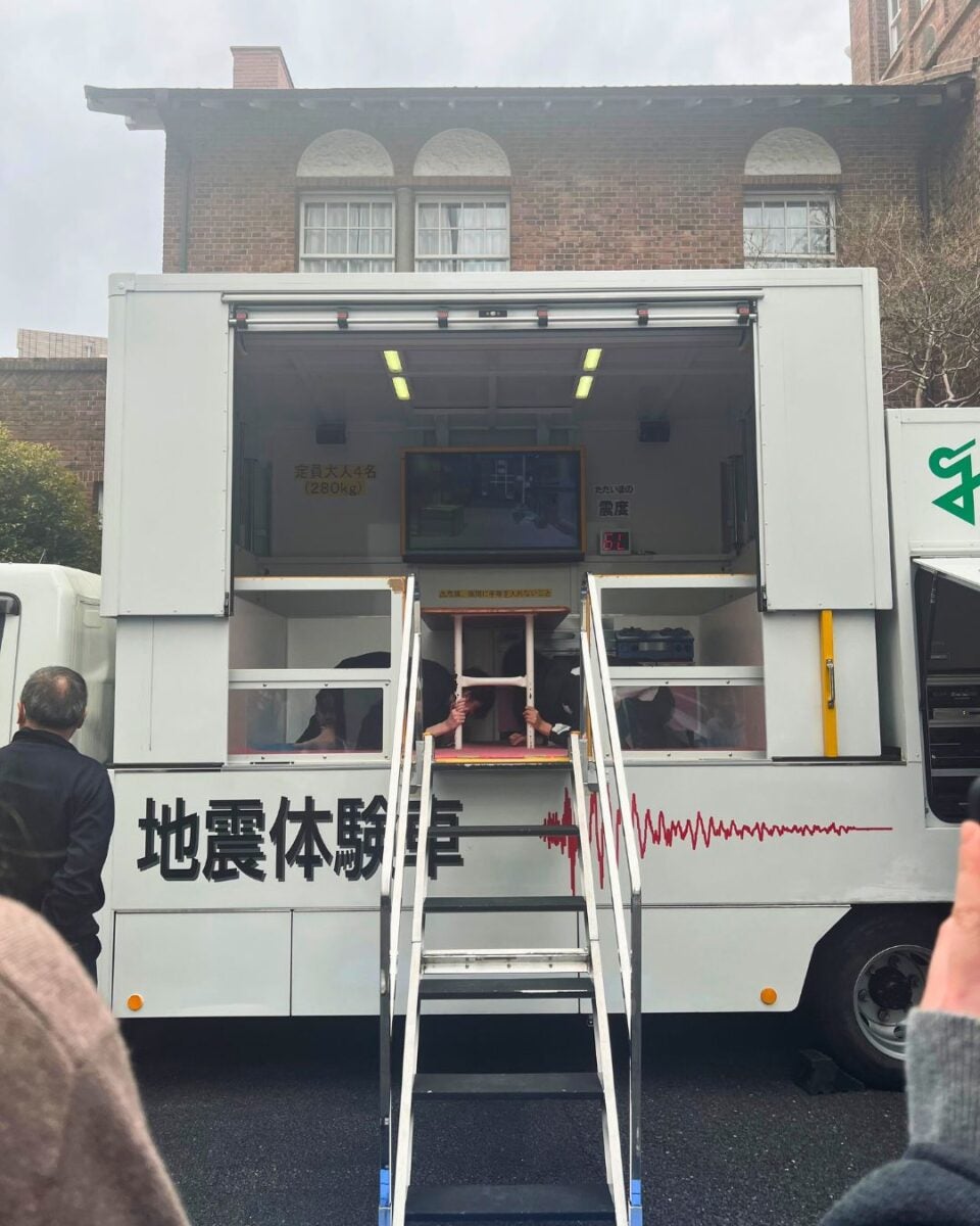 A white truck with open sides labeled with seismic waves in red, showcasing an earthquake simulator. Stairs lead up to a viewing area with people inside. The setting is in front of a brick building, and there's a cloudy sky.