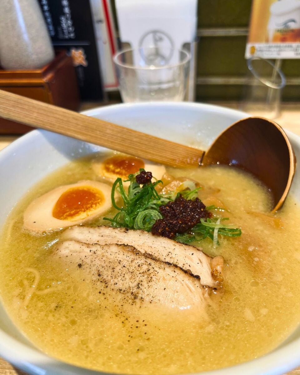 A bowl of ramen with slices of tender pork, soft-boiled eggs, green onions, and chili paste, immersed in rich, creamy broth; a wooden spoon rests on the bowl's edge.