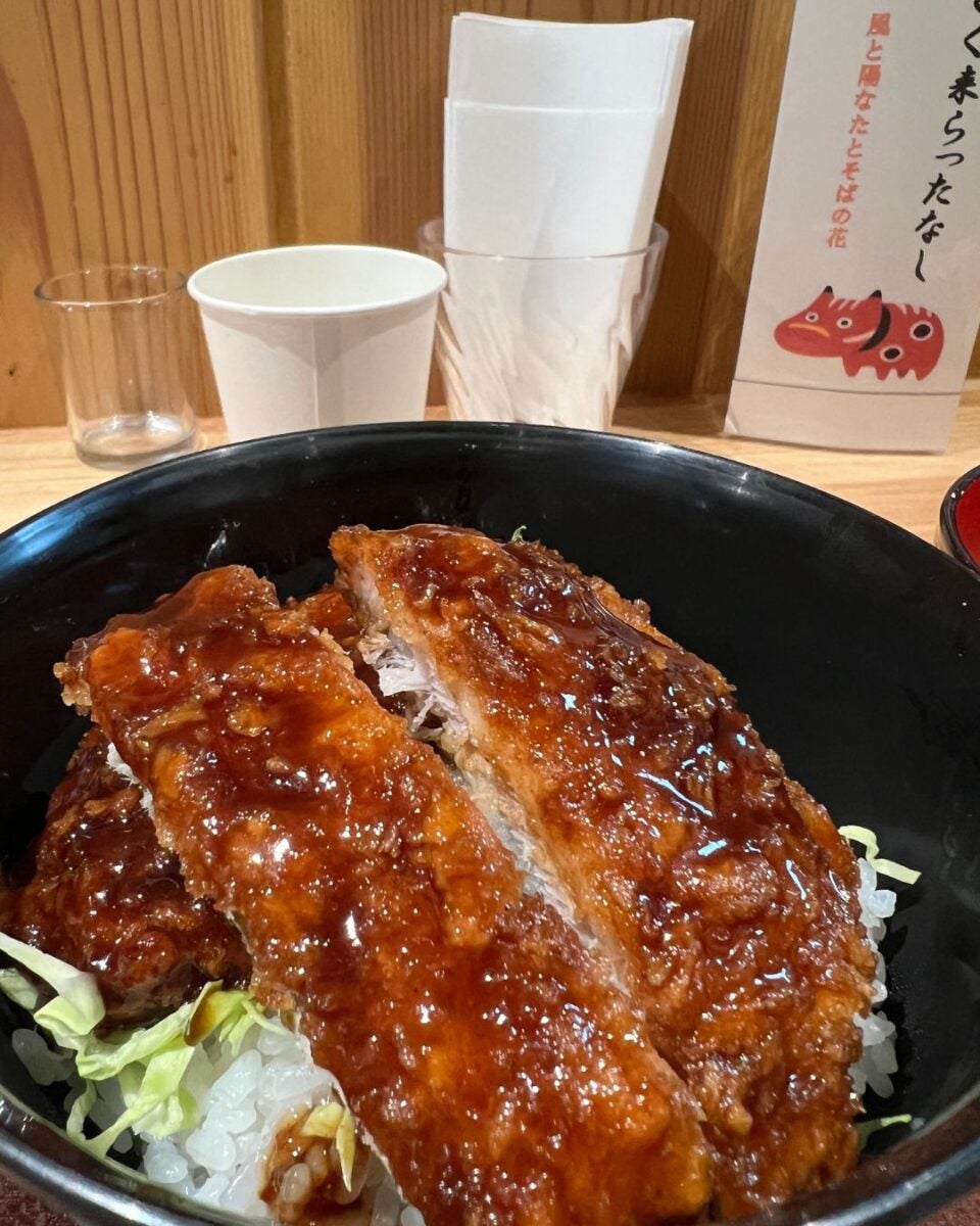 A black bowl of glazed katsu on rice with cabbage. Background shows a wooden wall with a cup, napkins, and a menu featuring a small, red animal illustration.