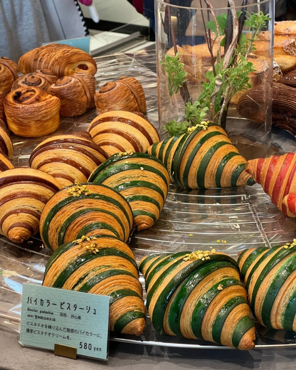 A display of croissants with green and brown stripes, resembling leaves. They are arranged on a tray near a small decorative plant. A price tag reads "580 yen."