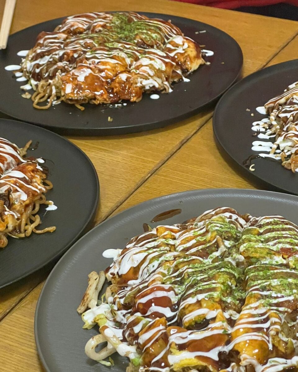 Plates of okonomiyaki on a wooden table. Each serving drizzled with mayo, okonomiyaki sauce, and green seaweed, creating a savory, appetizing appearance.