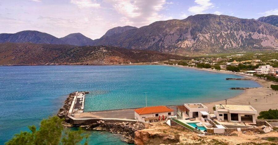 Coastal bay with a pier, modern buildings, a sandy beach, and rugged mountains in the background.