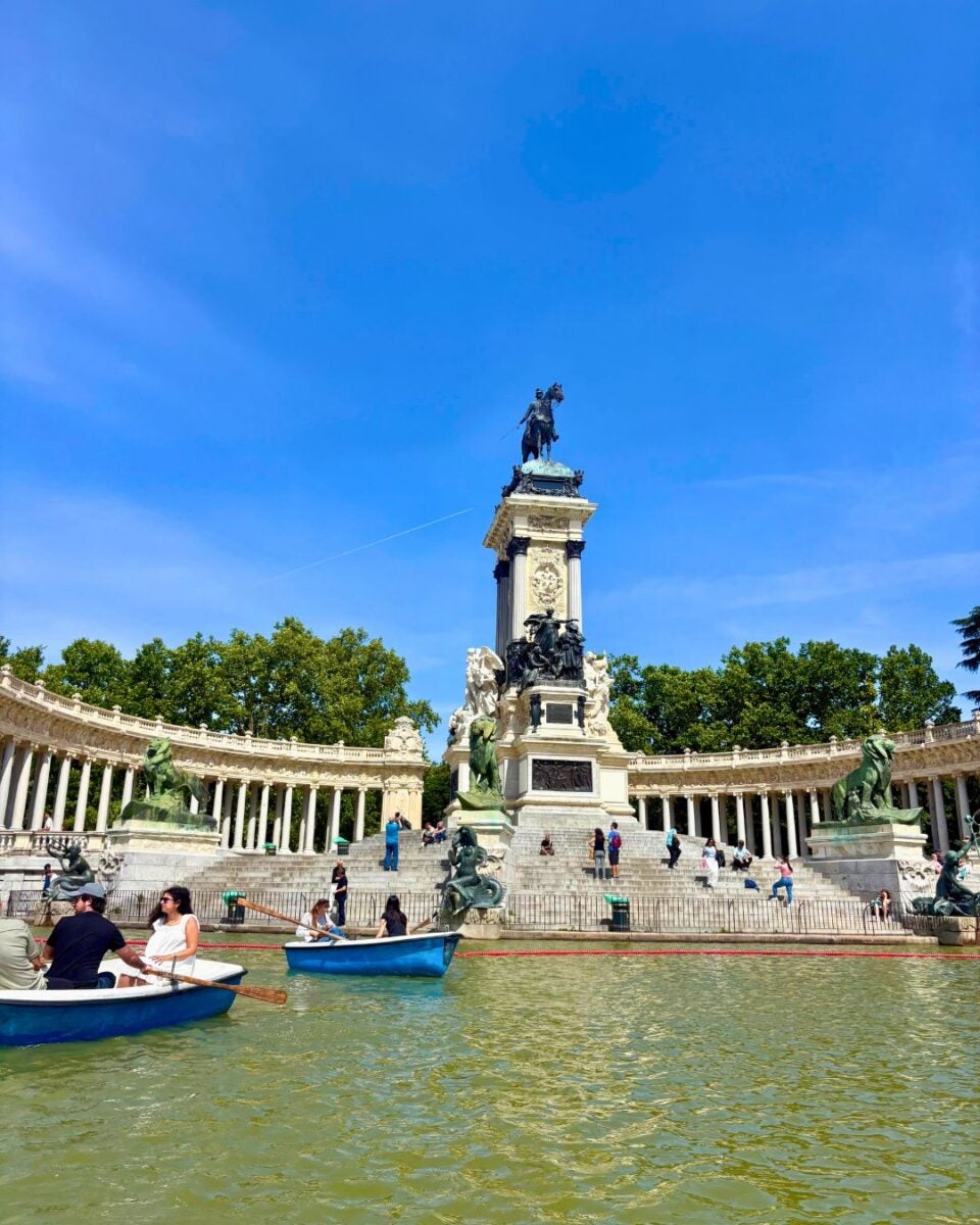 Monument surrounded by a semicircular colonnade and boats on a lake on a sunny day.