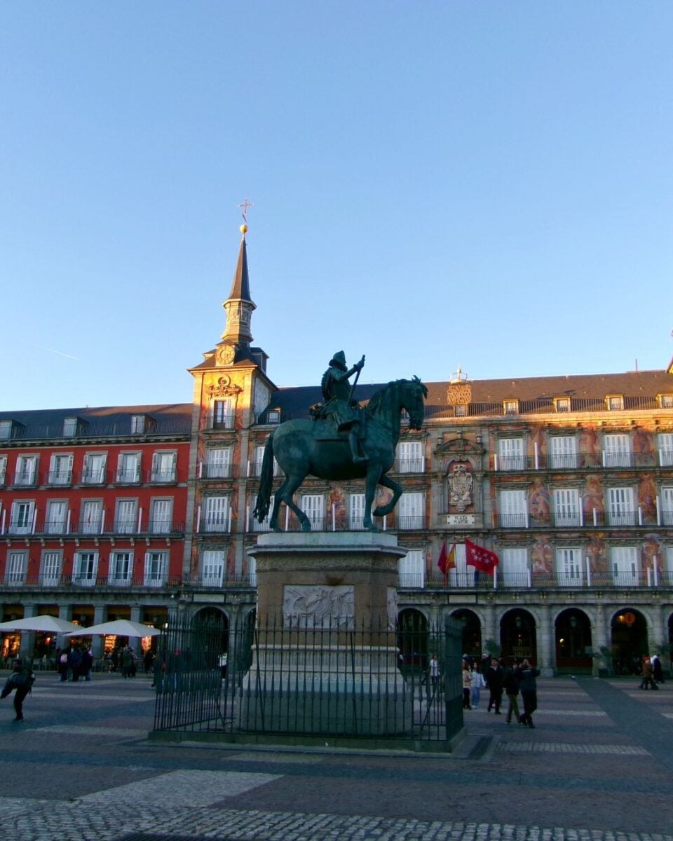 The image depicts the Plaza Mayor in Madrid, featuring a prominent bronze equestrian statue in the center, mounted on a stone pedestal.