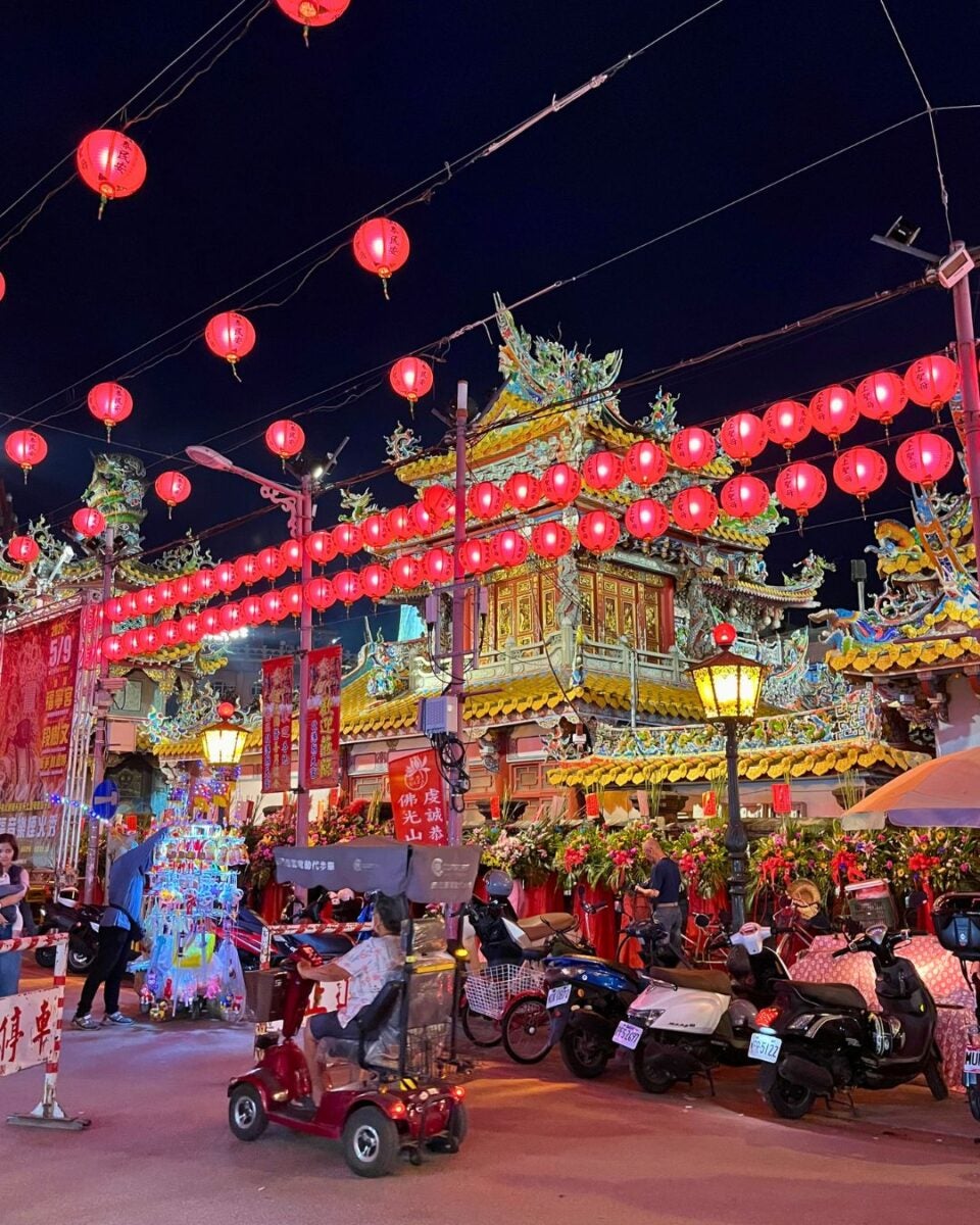A vibrant night scene at a Chinese temple, adorned with rows of glowing red lanterns. People and scooters are in the foreground, creating a festive atmosphere.