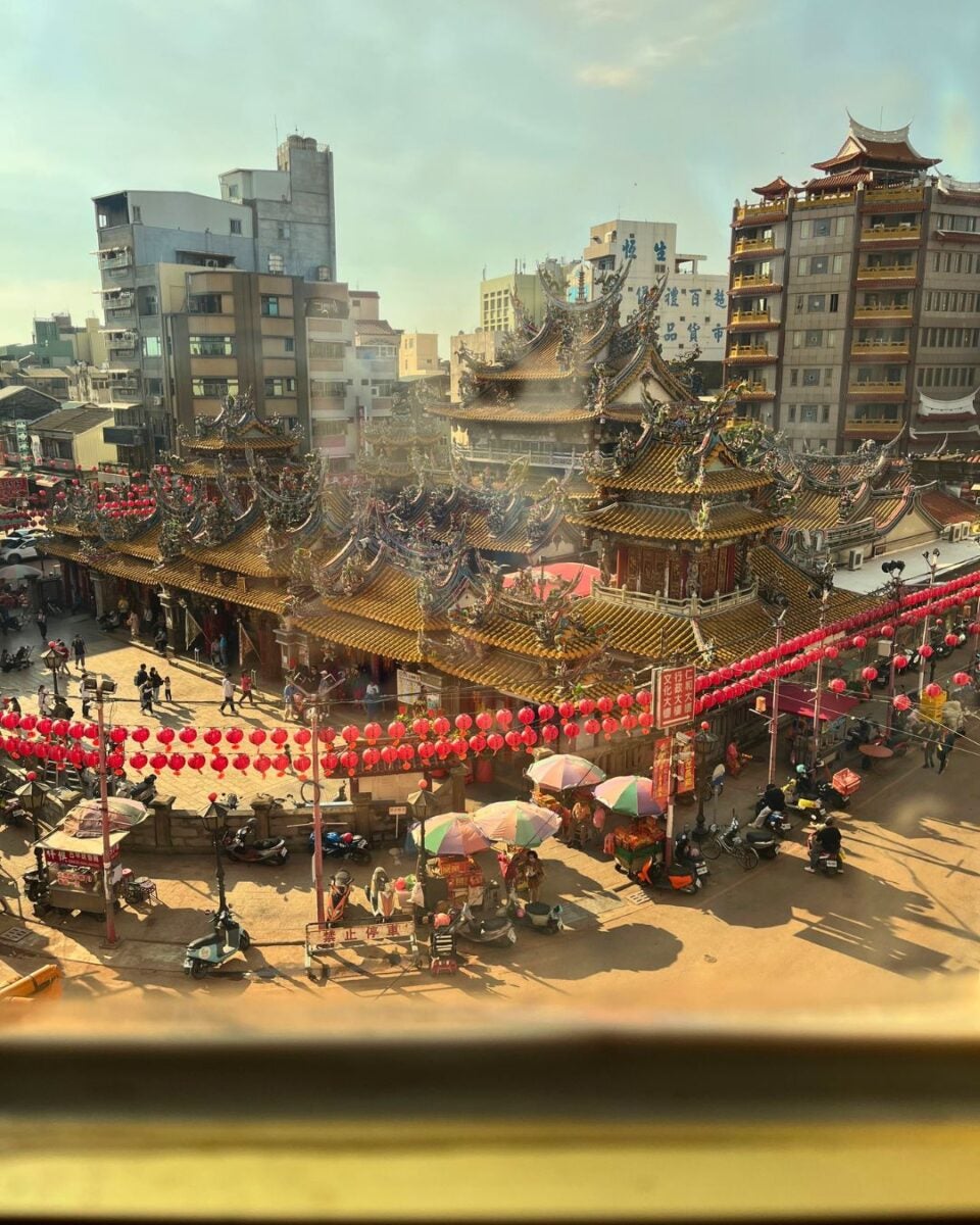 Rooftop view of a traditional Chinese temple with ornate details, surrounded by vibrant red lanterns. Street stalls and scooters line the bustling street below.