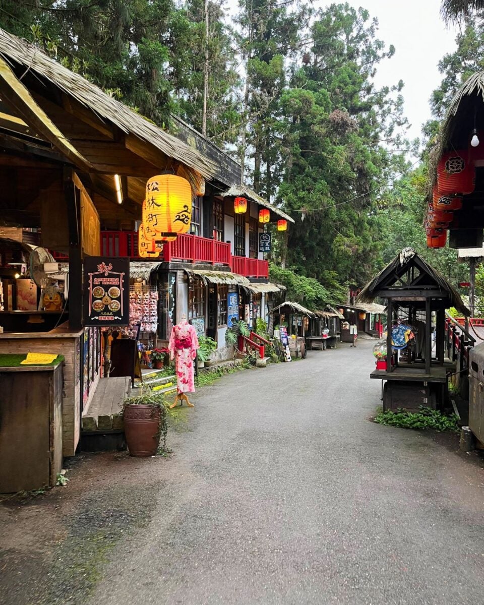 Charming village street lined with wooden shops and red lanterns, amidst lush trees. Traditional attire and signs add a nostalgic, serene vibe.