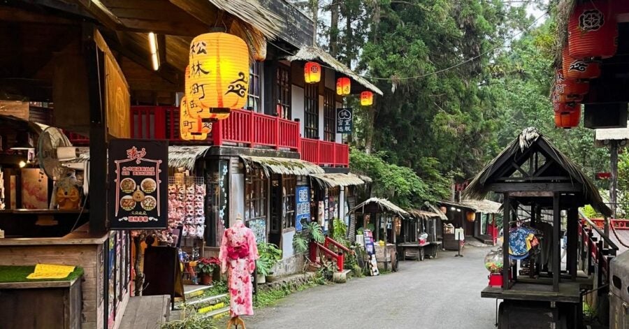 Charming village street lined with wooden shops and red lanterns, amidst lush trees. Traditional attire and signs add a nostalgic, serene vibe.