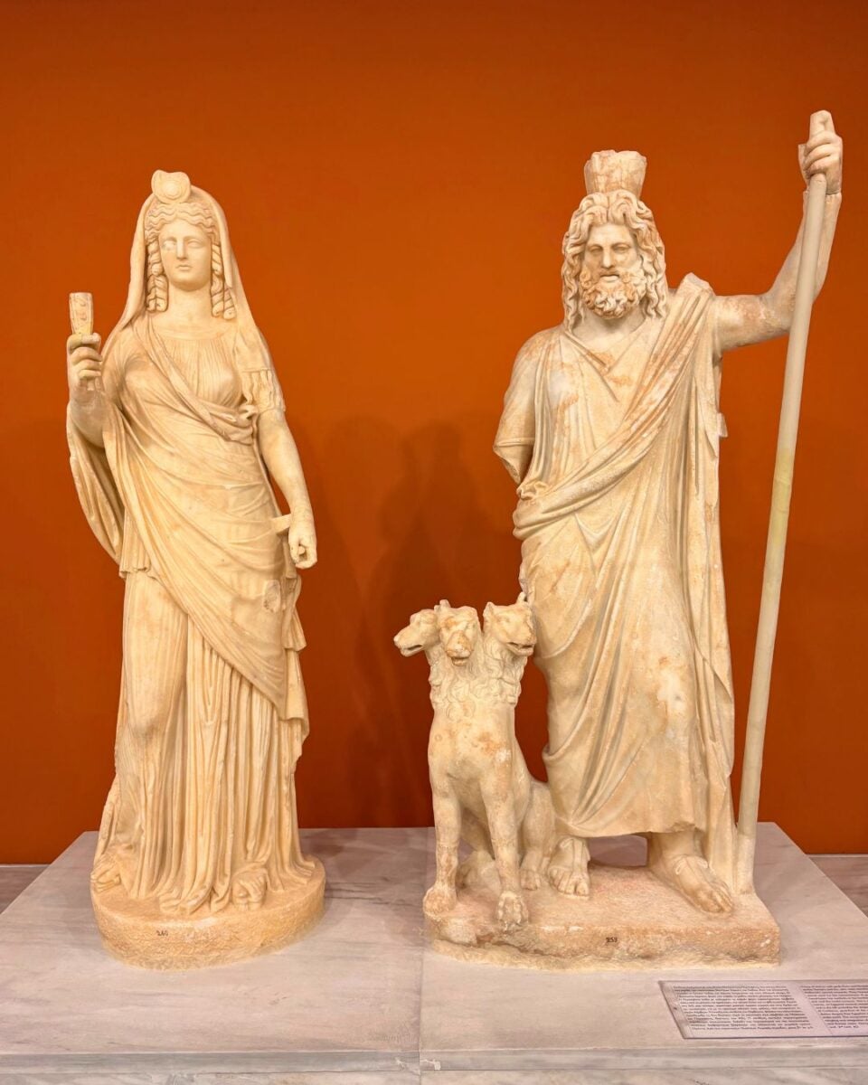 Two marble statues of a female and male figure with a three-headed dog, set against an orange background.