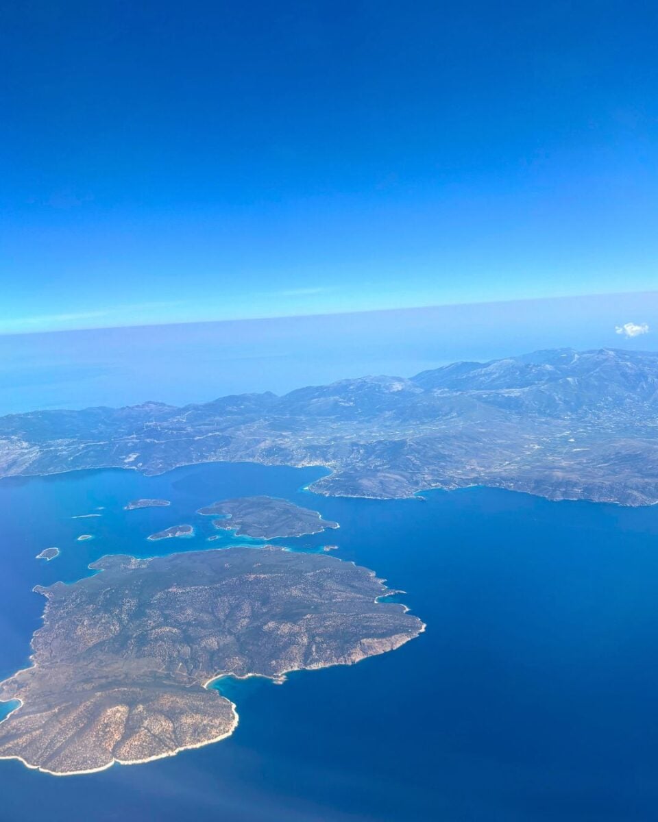 Aerial view of islands with green vegetation and sandy beaches surrounded by blue ocean water, with mountains and clear sky in the background.