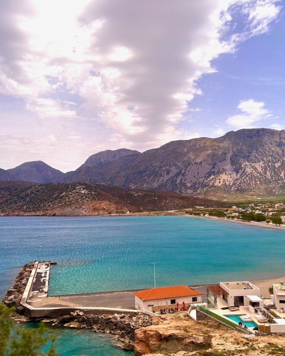 Coastal bay with a pier, modern buildings, a sandy beach, and rugged mountains in the background.