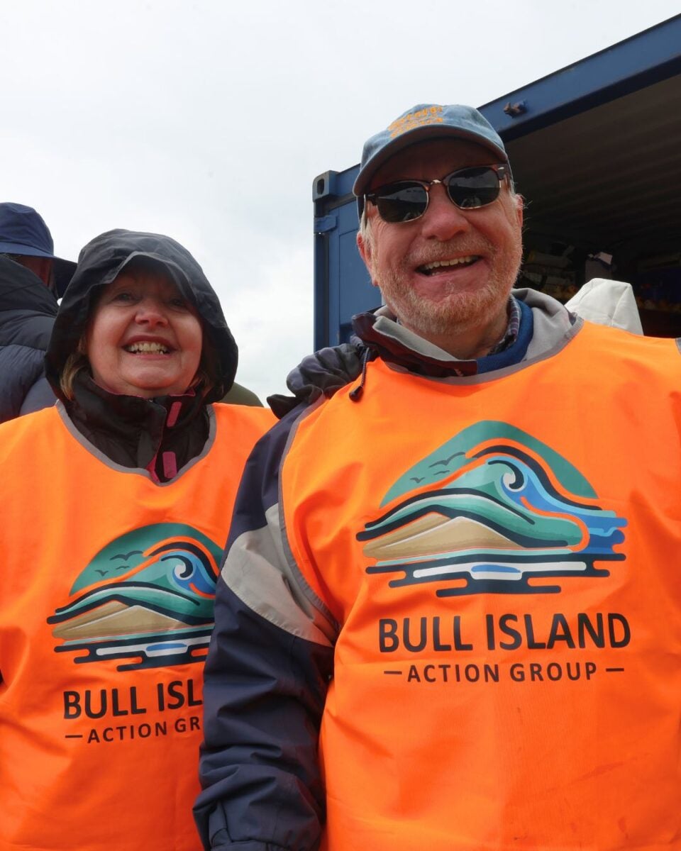 A woman and a man in orange vests with a coastal logo, smiling outdoors under an overcast sky.