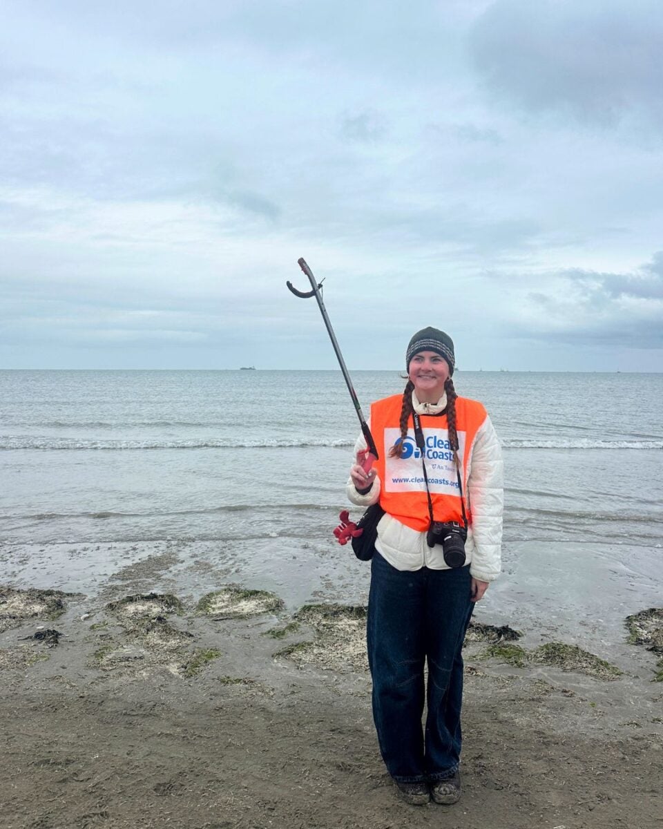 A woman on a beach holding a litter picker, wearing an orange safety vest.