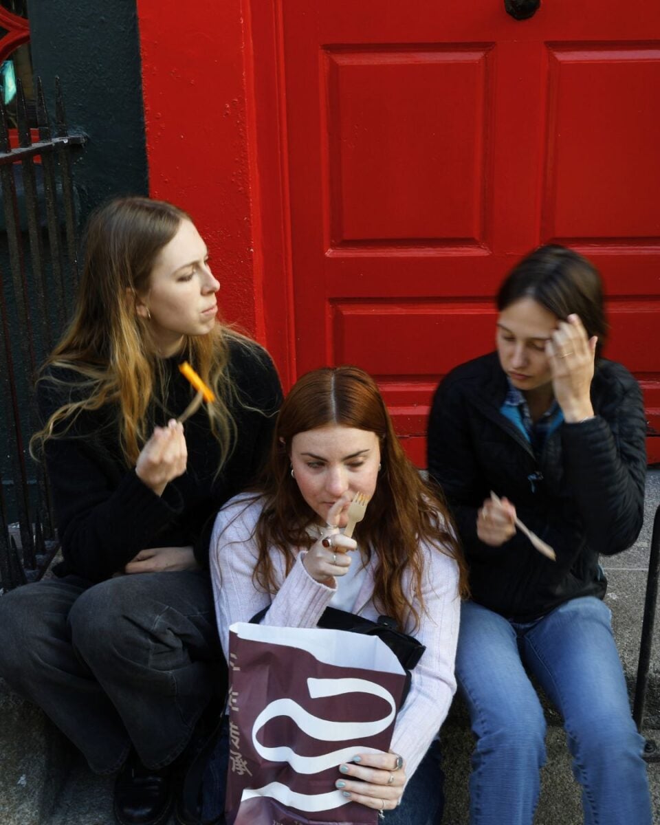 Three young women sitting on steps in front of a red door, eating from a bag.