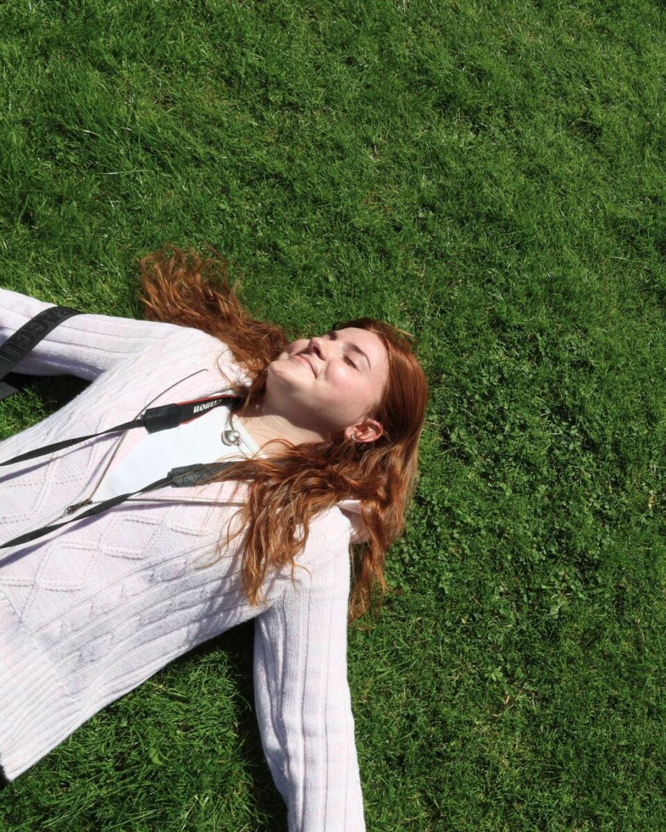 A woman with red hair lying on green grass, wearing a white sweater.