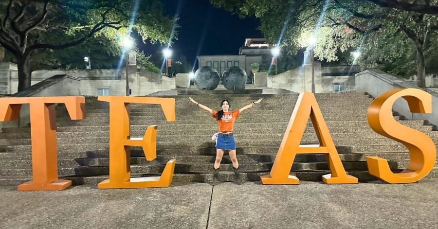 A person joyfully stands with arms raised between large, illuminated orange letters spelling "TEXAS" on stone steps at night, under trees and lampposts.