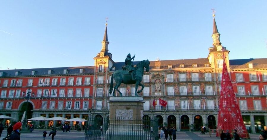 The image depicts the Plaza Mayor in Madrid, featuring a prominent bronze equestrian statue in the center, mounted on a stone pedestal.