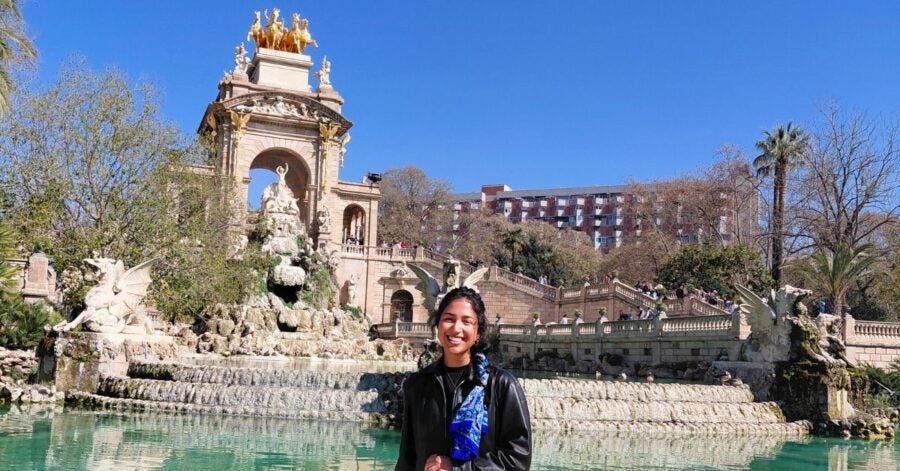 A person smiles in front of a grand fountain with ornate sculptures and cascading water. The sky is clear and blue, creating a vibrant, joyful atmosphere.