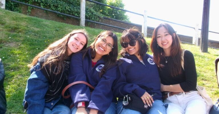 Four young women sitting together on grass, smiling on a sunny day.