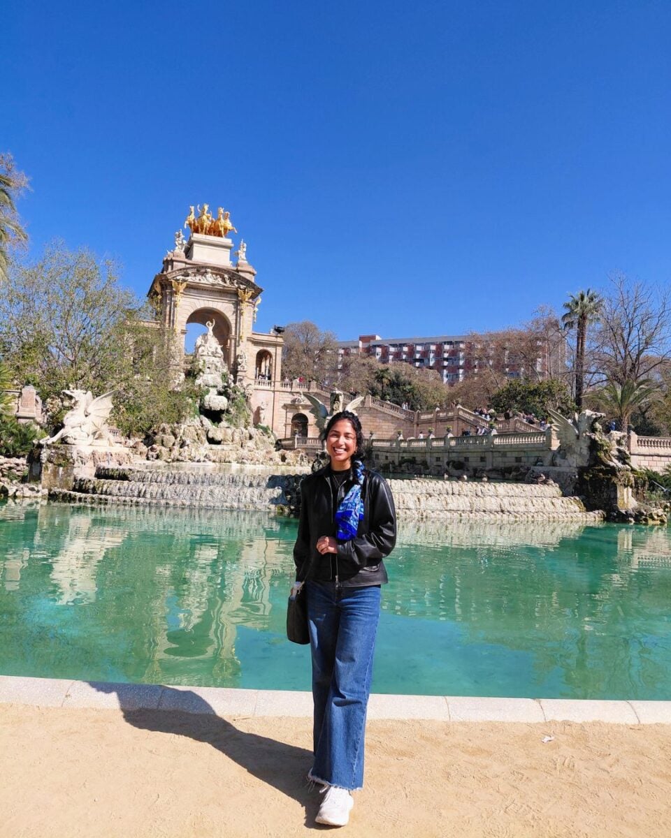 A person smiles in front of a grand fountain with ornate sculptures and cascading water. The sky is clear and blue, creating a vibrant, joyful atmosphere.