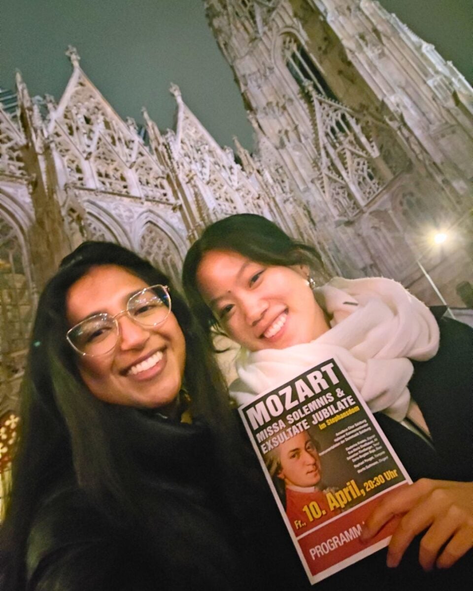 Two smiling people pose at night in front of a Gothic cathedral, one holding a Mozart concert flyer. The mood is joyful and enthusiastic.