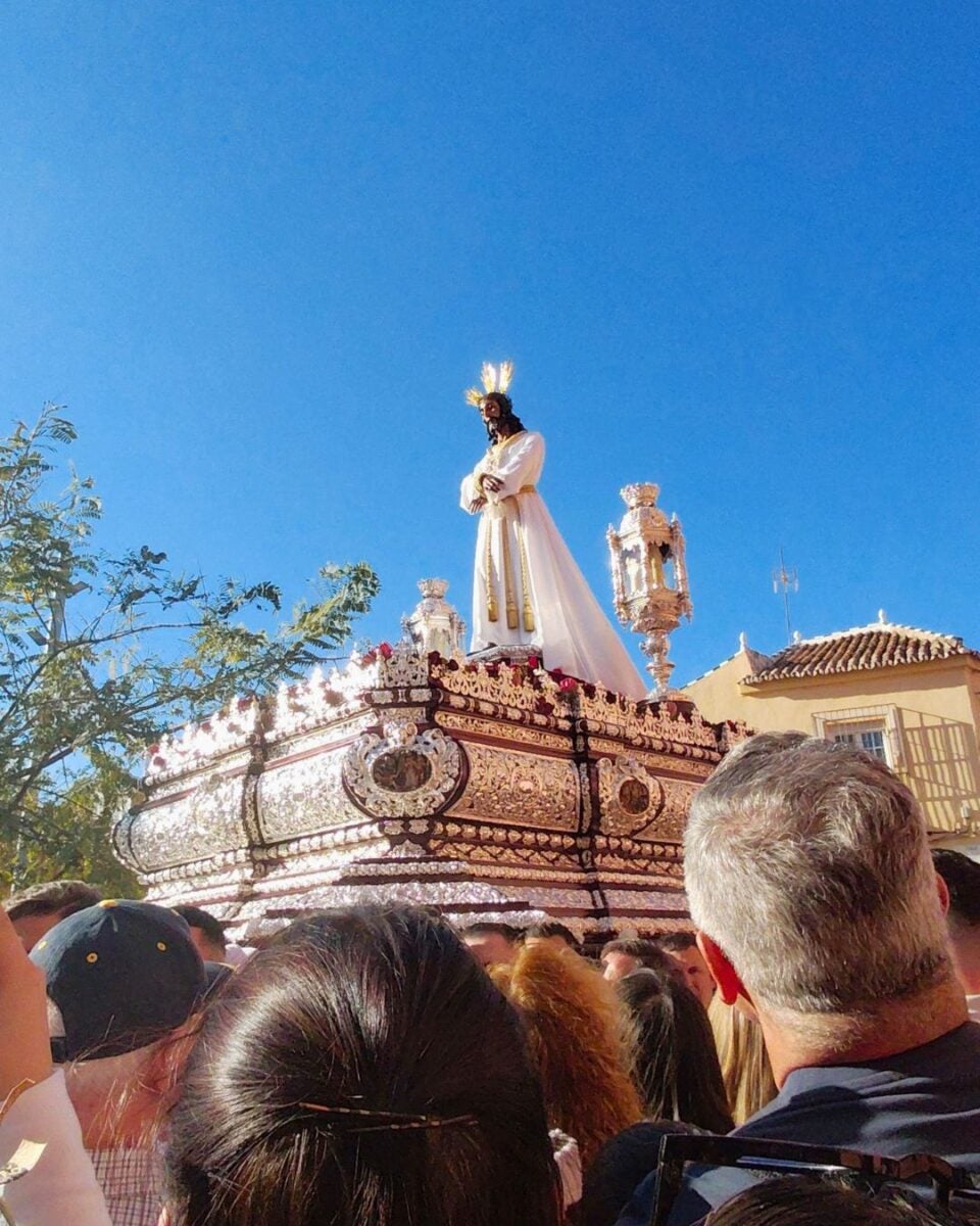 A religious procession features a statue of Jesus in white robes on a lavishly decorated float. A clear blue sky and a large crowd are in the background.