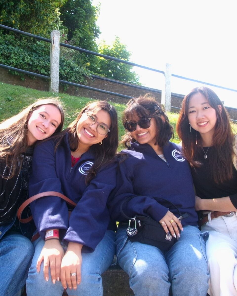 Four young women sitting together on grass, smiling on a sunny day.
