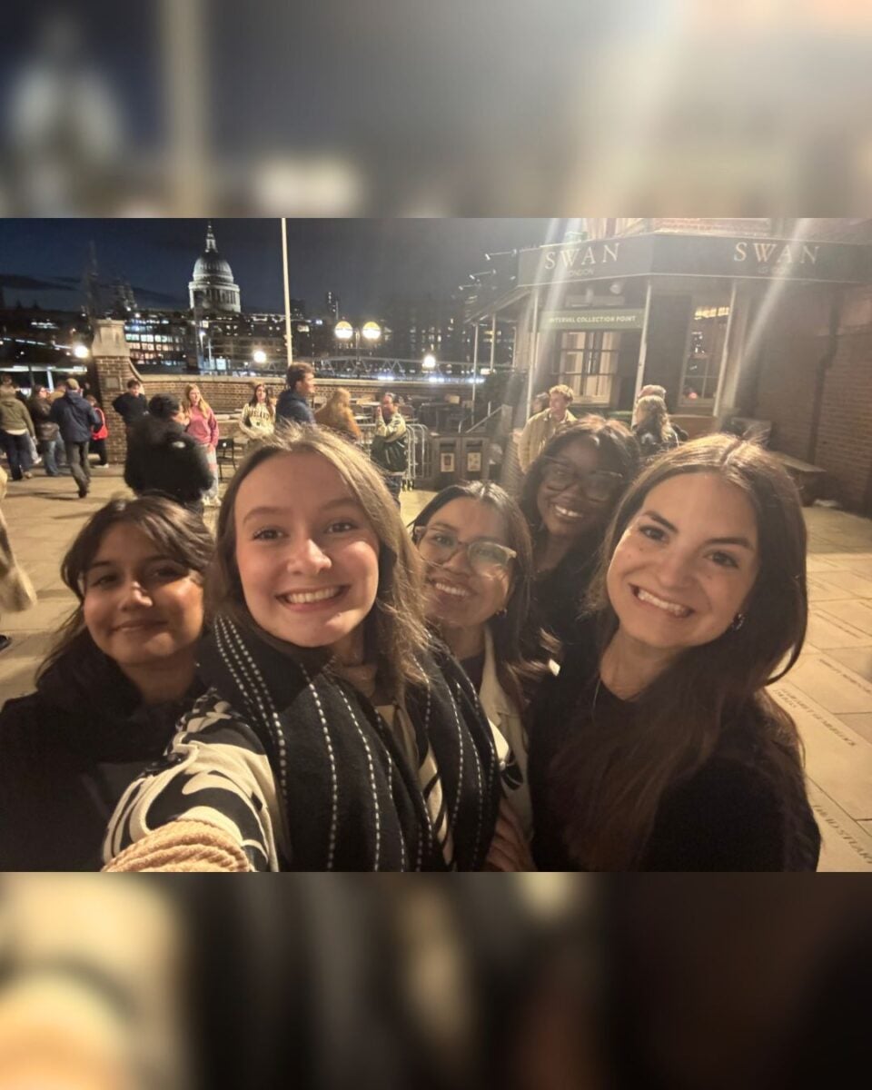 A group of five young women smiling at the camera with a cityscape and dome-shaped building in the background at night.