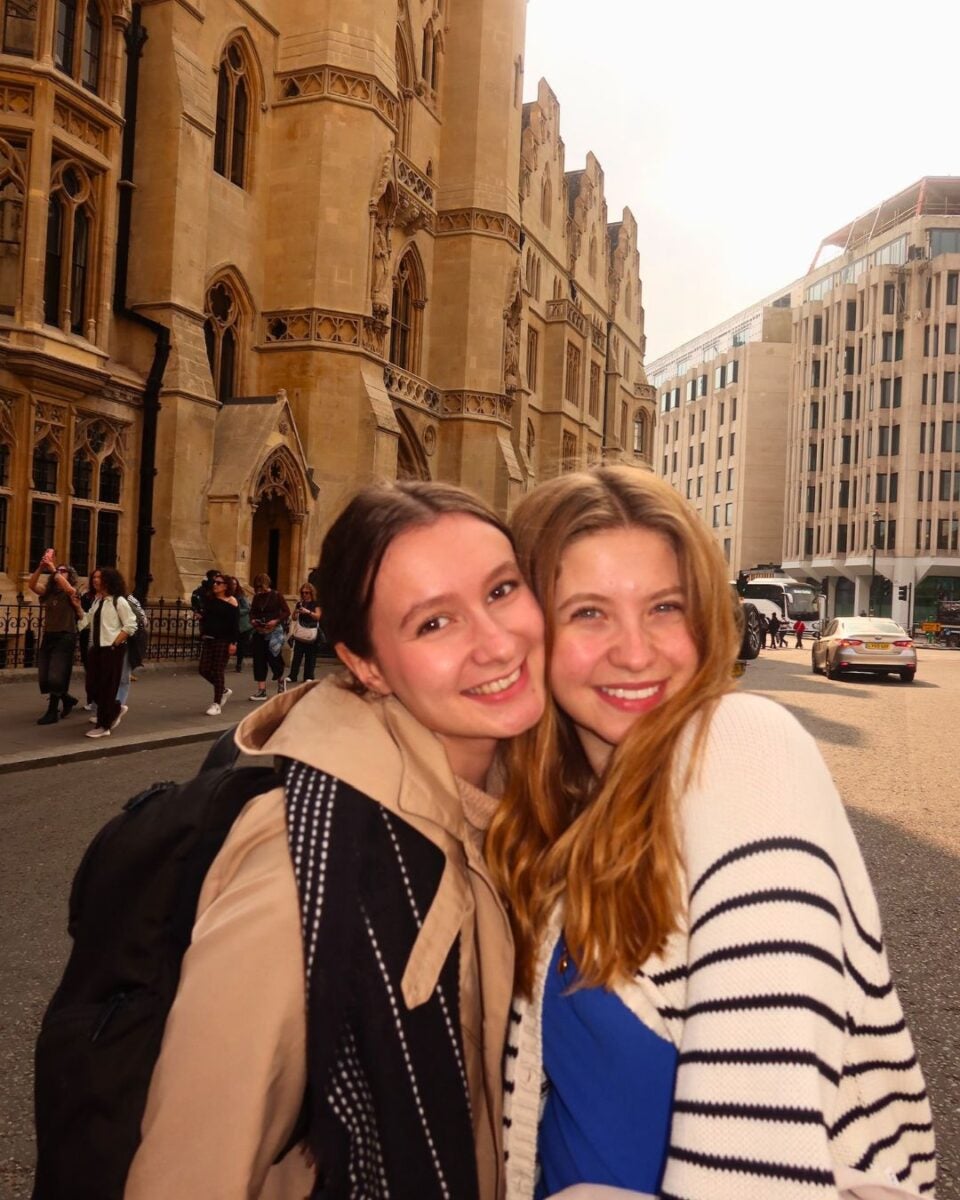 Two young women smiling in front of Gothic-style and modern buildings on a city street.