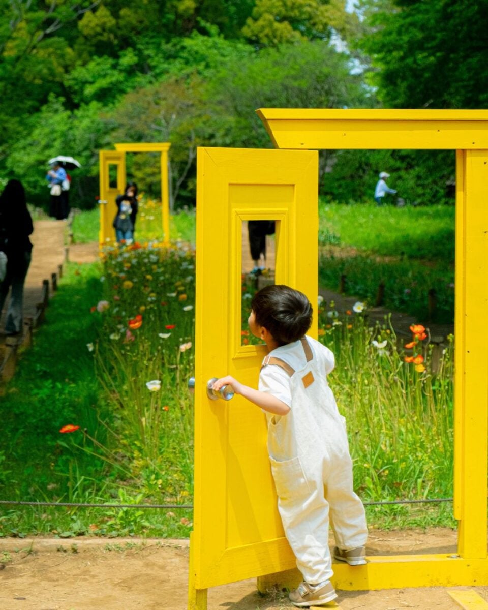 A child in white overalls reaches for a bright yellow door in a garden. Wildflowers dot the path, and more yellow doors stand in the lush greenery beyond.