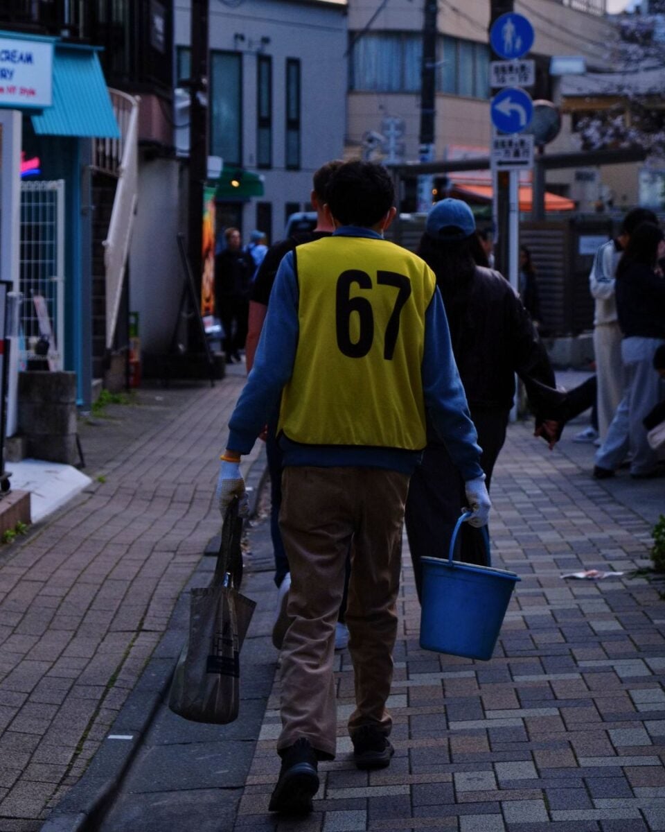A person wearing a yellow vest with number 67 walks down a dimly lit street, carrying a blue bucket and a trash bag. They appear engaged in cleaning work.