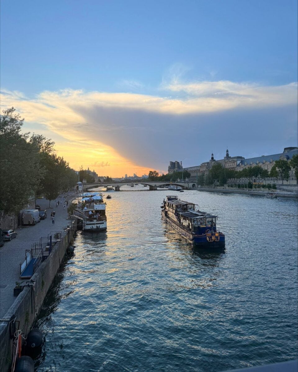 Sunset over a river with boats cruising and a bridge in the distance under a partly cloudy sky.
