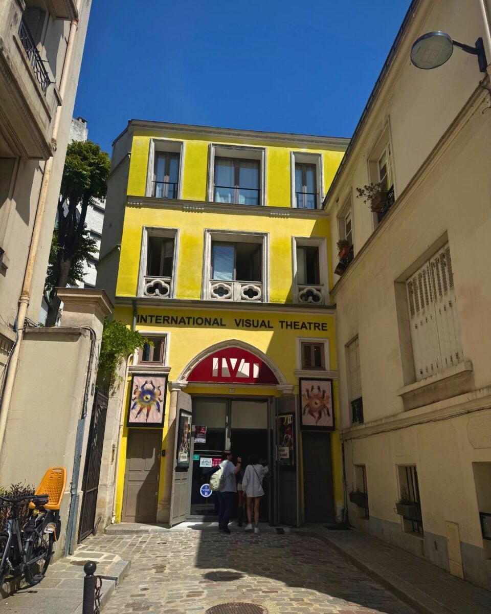 Bright yellow facade of the International Visual Theatre with open entrance and two people standing outside under a clear blue sky.