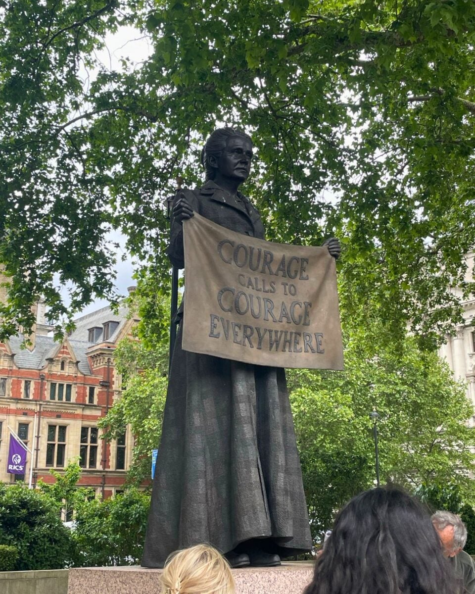 A statue of a person holding a banner that reads, "Courage calls to courage everywhere," surrounded by trees and an ornate building in the background.