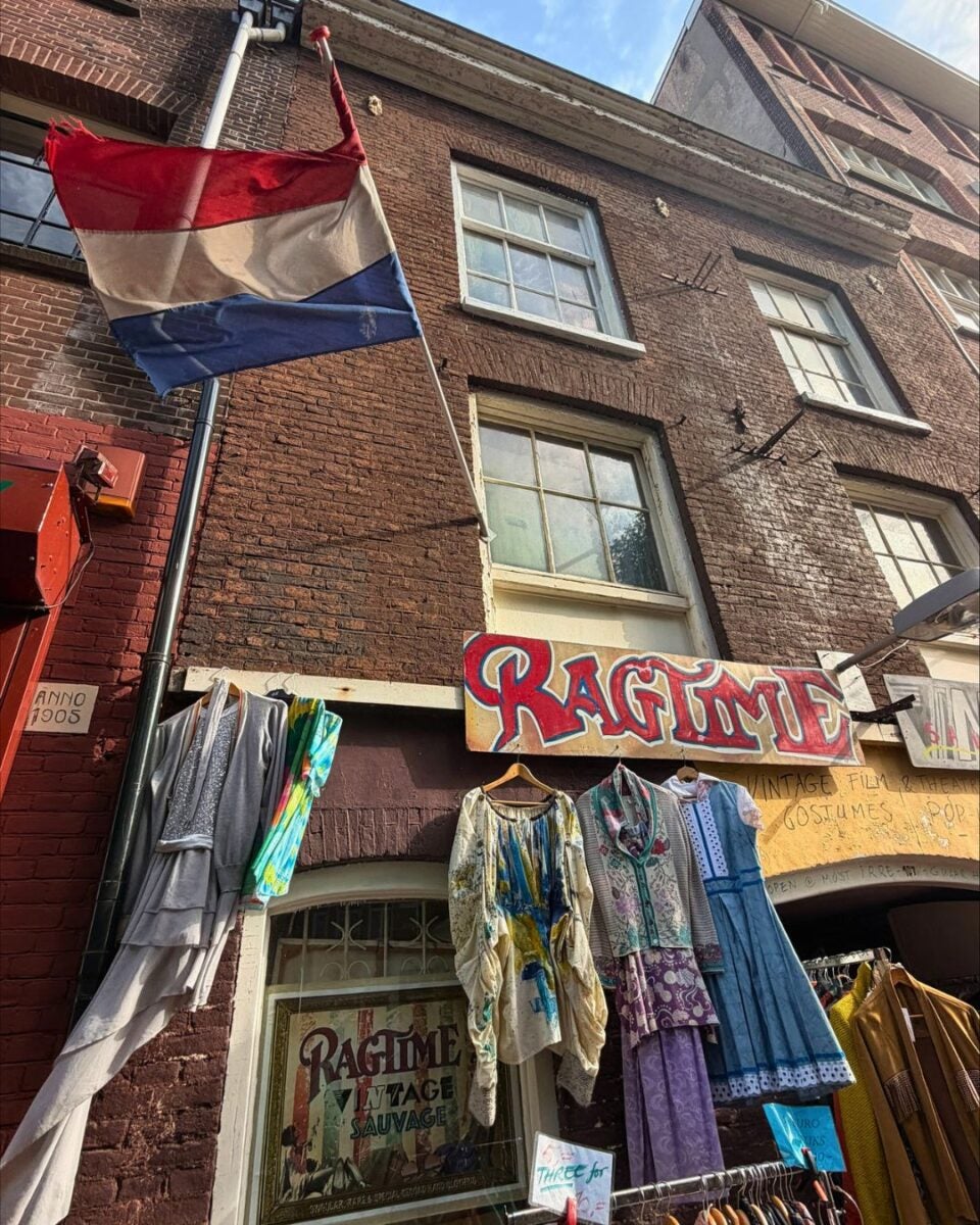Dutch flag flying above a vintage clothing shop named Ragtime with colorful dresses displayed outside on hangers.
