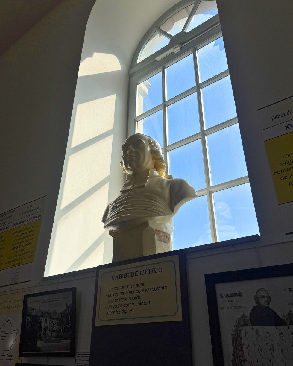 Bust of a historical figure illuminated by sunlight in front of a large arched window with blue sky outside.