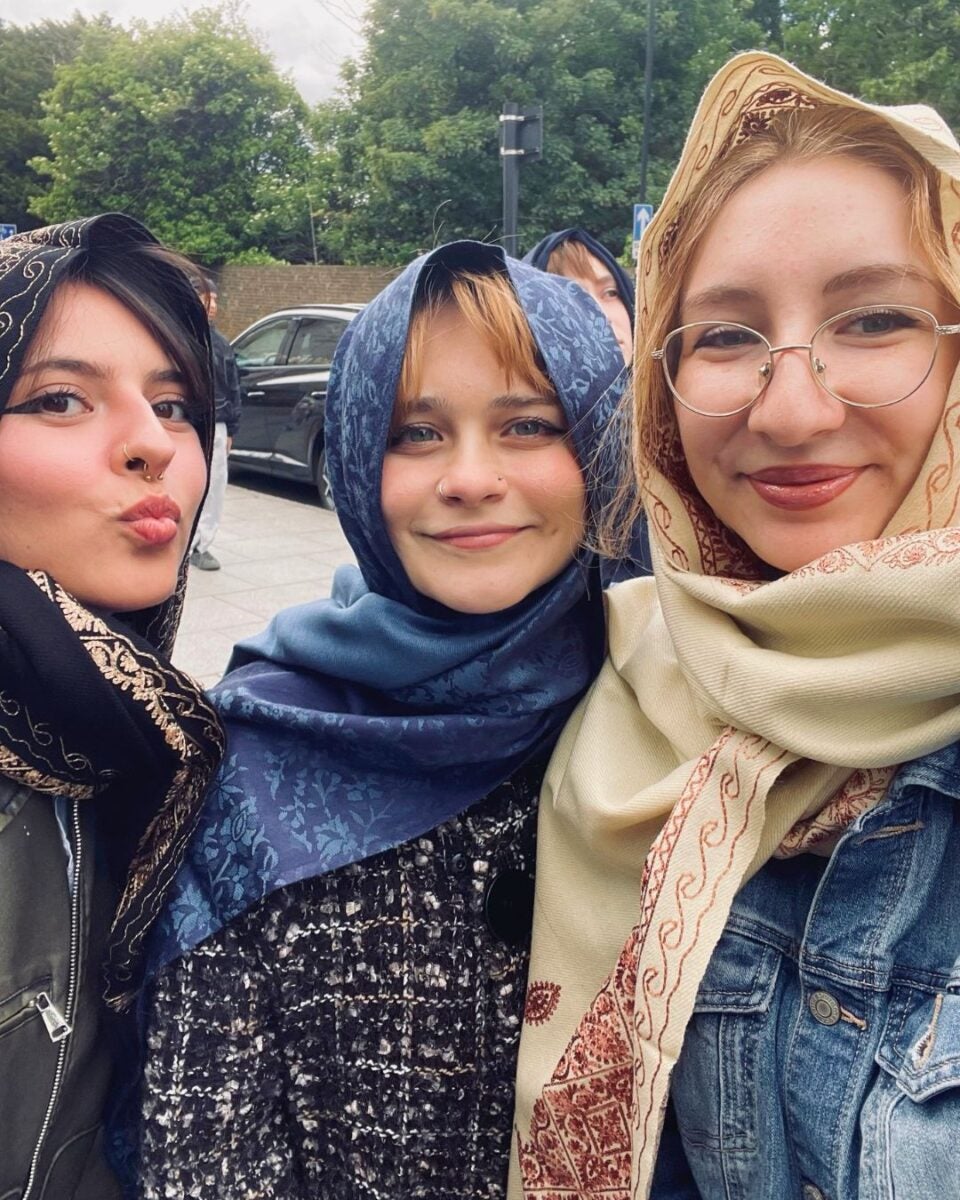 Three young woman with headscarves, standing close together, with a park and car in the background.
