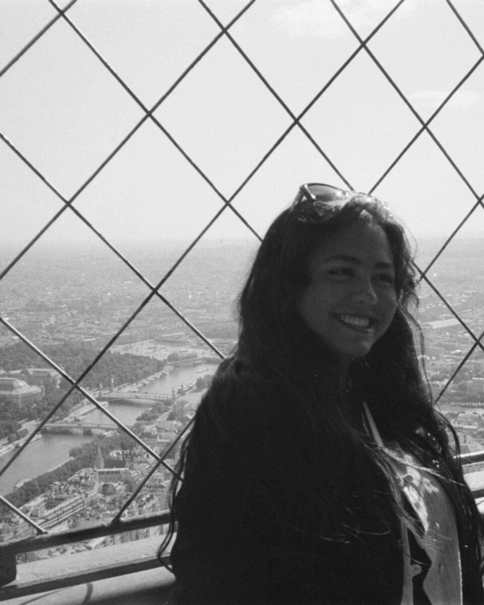 A young woman with long hair standing behind a safety net on a high observation deck overlooking a cityscape with rivers and buildings in the distance.