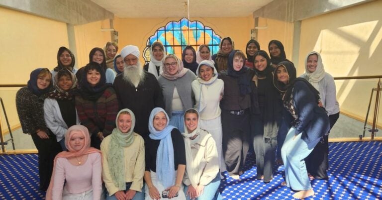 A group of people, mostly women wearing headscarves, pose together and smile for a photo inside a bright room with blue patterned carpet and arched stained glass.