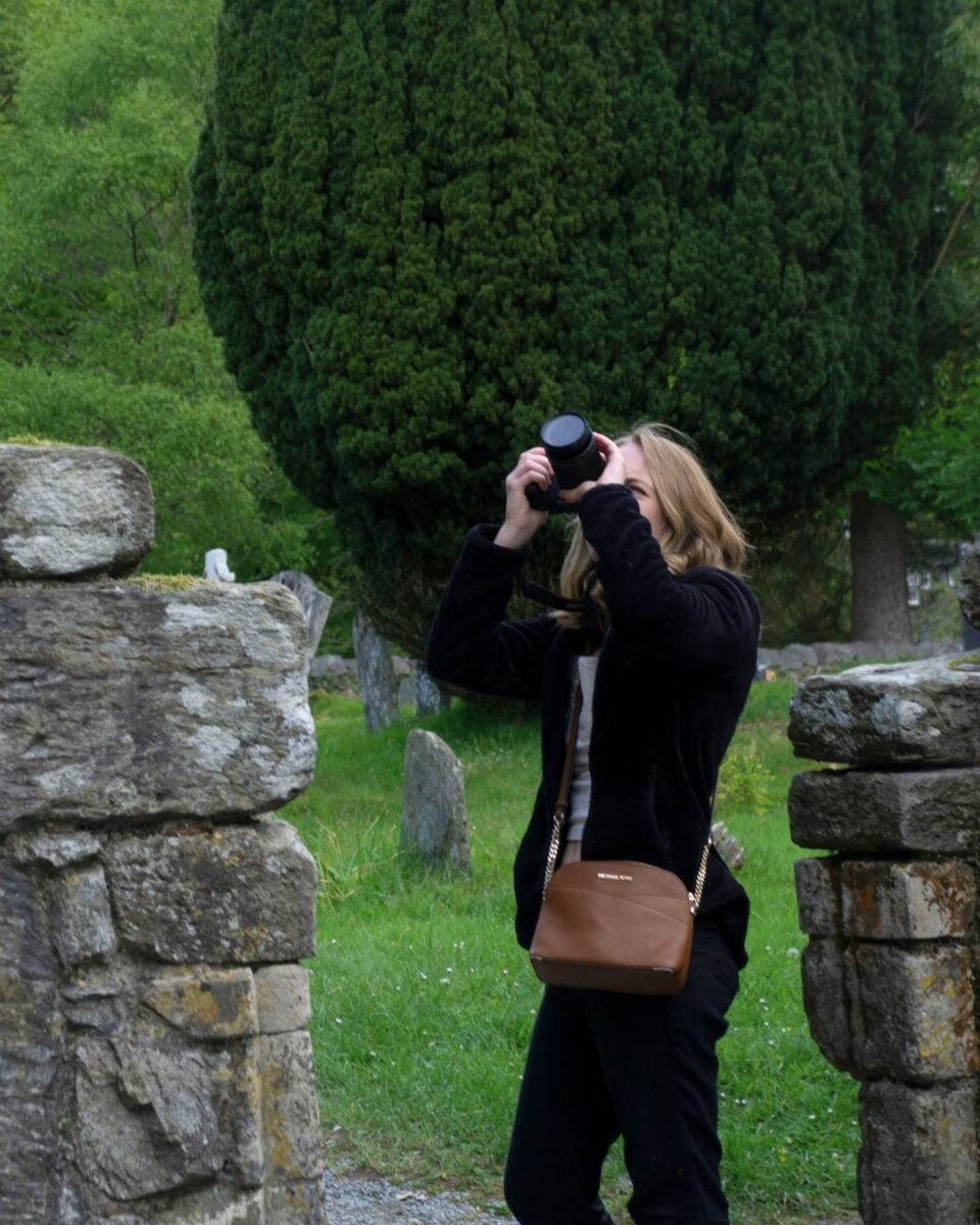 Woman in black jacket taking a photo with a camera in a green park surrounded by stone ruins and tall trees.